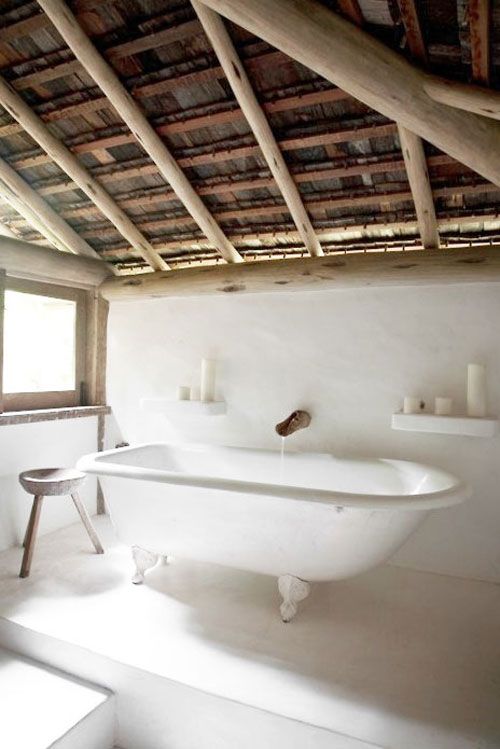 An airy white bathroom with a wooden beam roof, a claw-foot tub, a stool and a few shelves with pillar candles looks like a spa