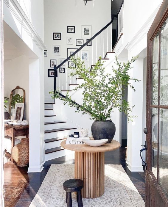 a chic and stylish entryway with a black and white gallery wall above the stairs, a wooden table with a large planter and green plants and a stool